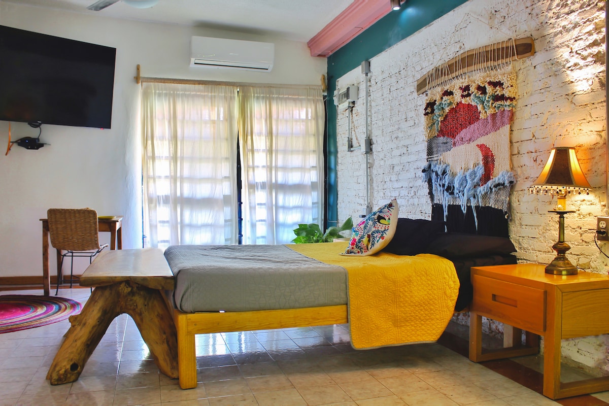 A spacious room features large windows covered by sheer curtains, allowing natural light to illuminate the interior. A wooden bed with a gray blanket contrasts against a textured brick wall. A decorative wall hanging adds color, while a small table and lamp provide additional lighting.