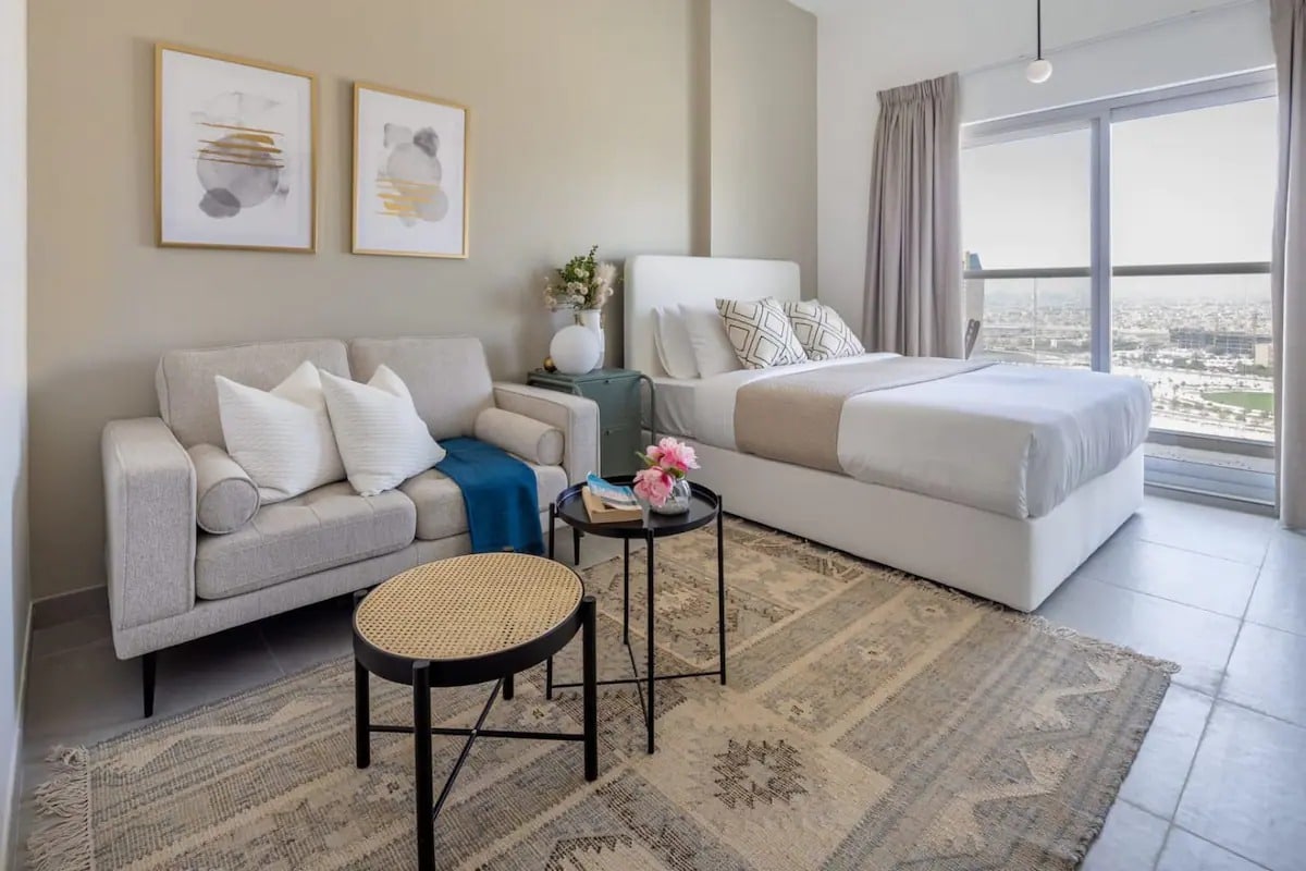 A spacious studio apartment features a comfortable sofa accompanied by decorative cushions and a minimalist coffee table. A king-sized bed with neutral linens is positioned against the wall, while large glass doors provide access to a balcony and showcase a city view.
