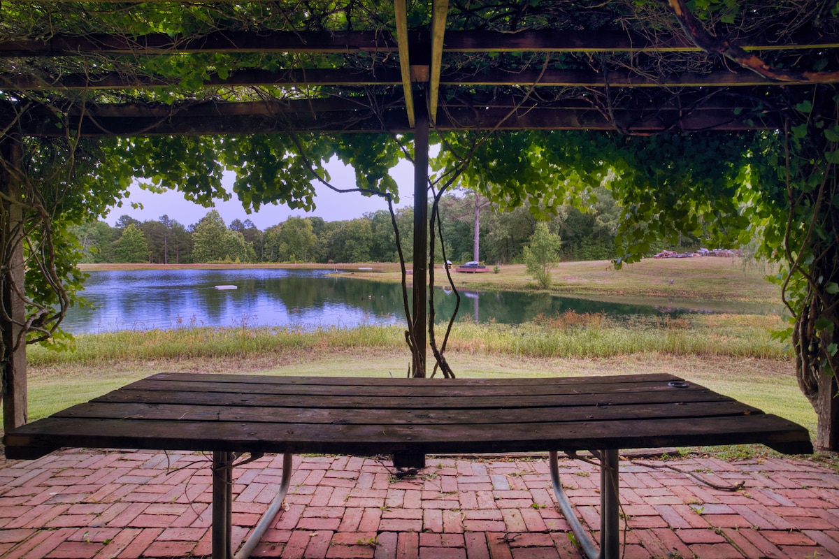 A shaded outdoor seating area is captured under a wooden trellis adorned with lush greenery. A rustic table sits atop a brick patio, offering a view of a tranquil pond that reflects the surrounding trees and open space.