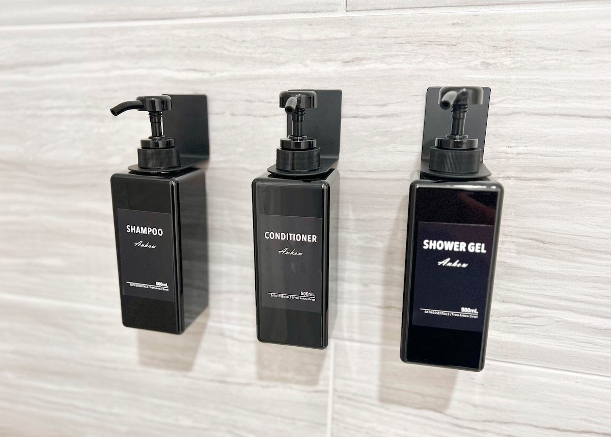 In a stylish bathroom, three pump bottles are mounted on a wall. The bottles are labeled 'Shampoo,' 'Conditioner,' and 'Shower Gel,' each designed with a sleek black finish. The contemporary tiles in the background add a modern touch to the space.