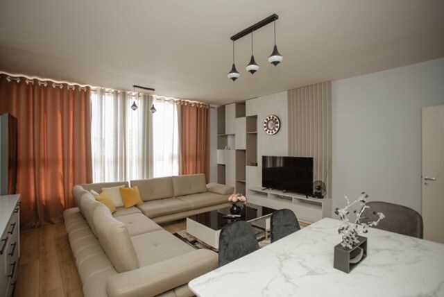 A spacious living room features a large sectional sofa and a modern coffee table. Natural light filters through floor-to-ceiling curtains, illuminating the space. A flat-screen TV is mounted on the wall, complemented by a minimalist shelving unit and a dining area with elegant chairs.