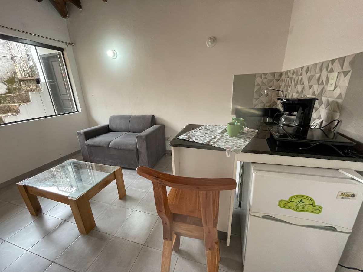 A bright kitchen and living area is showcased, featuring a small dining table and a comfortable gray sofa. The kitchenette includes a refrigerator and coffee maker. A large window allows natural light to enhance the space, while simple decor adds to the overall functionality.