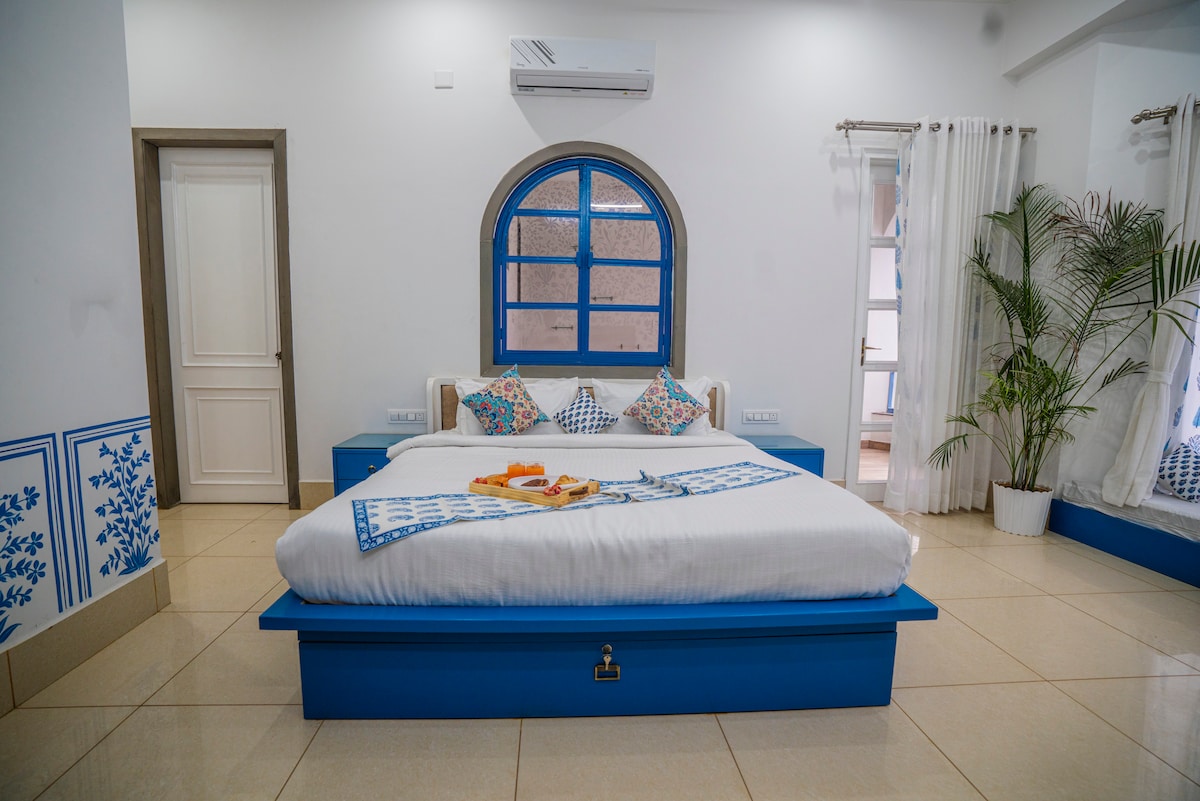 A spacious bedroom features a large bed with a crisp white comforter and colorful accent pillows. Blue cabinetry complements the warm tile flooring, while natural light filters through white curtains at the window. Decorative plants add a touch of greenery to the inviting space.