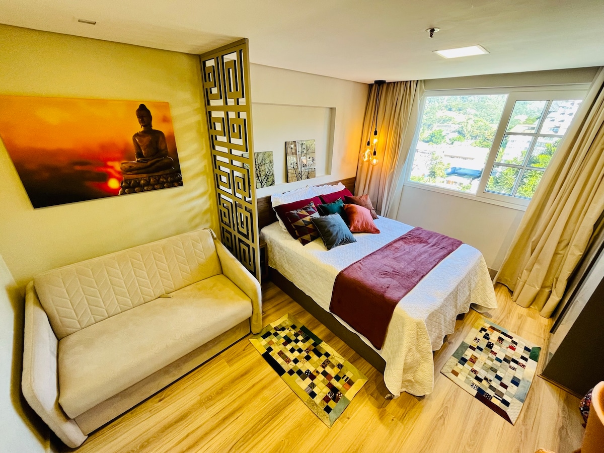 The room features a spacious bed adorned with a red and white bedspread, complemented by colorful pillows. A cozy sofa is positioned nearby. A serene Buddha artwork hangs above the bed, and two small rugs add warmth to the modern wooden floor. Natural light floods in through the window.