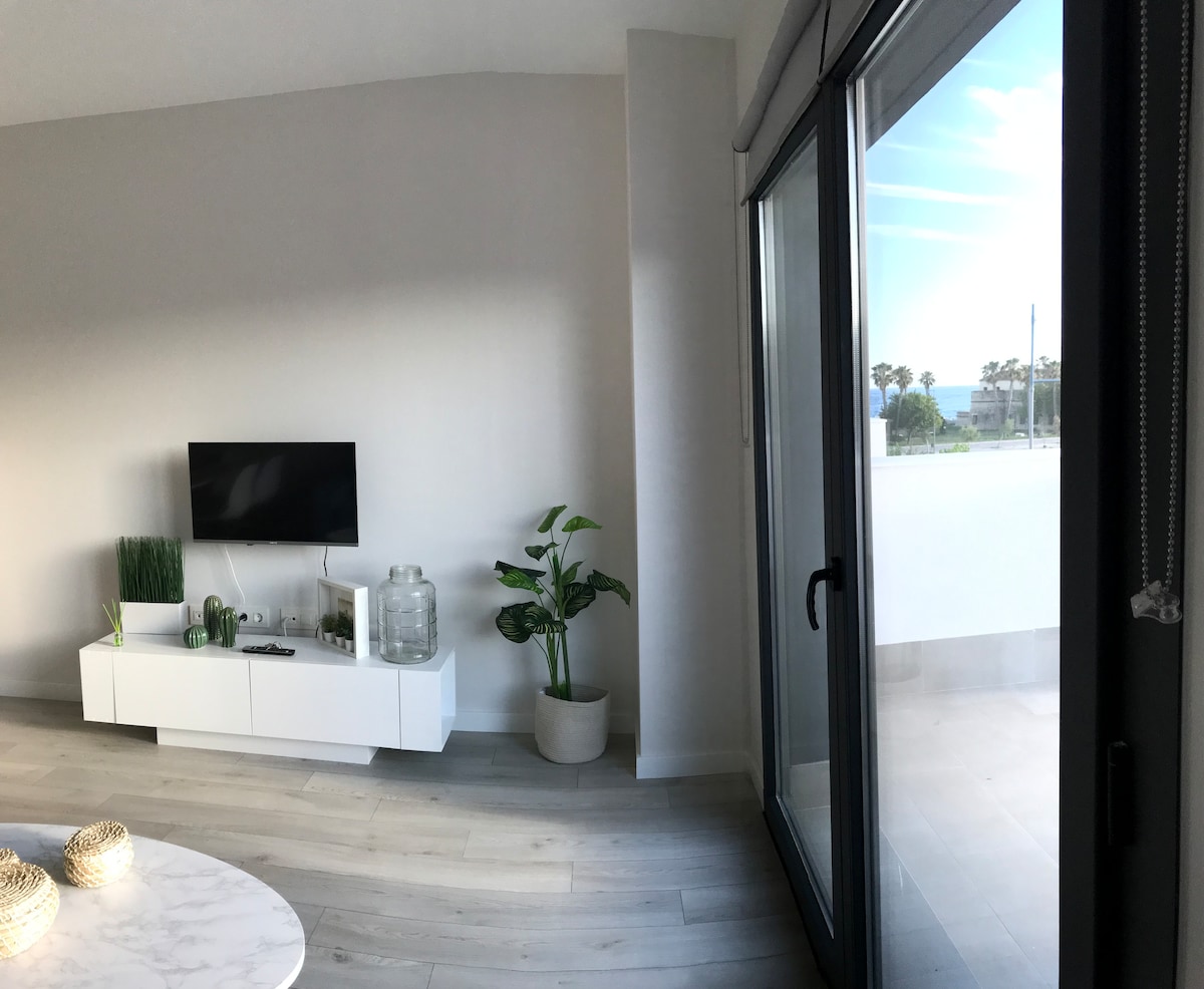 A modern living space is highlighted by a white entertainment unit housing a television and decorative plants. Large glass doors open to a private outdoor area, providing glimpses of the sea and embracing natural light within the bright, neutral-toned interior.