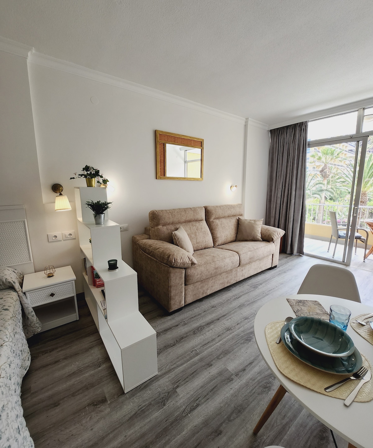 A living area is displayed with a comfortable sofa positioned near a glass door that leads to a balcony. A round dining table set is visible, accompanied by a light-colored shelving unit. Natural light fills the space, enhancing the layout.