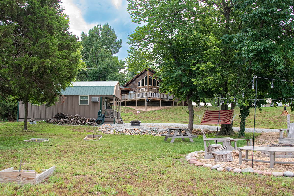 A spacious outdoor area features both a rustic cabin and a larger home, surrounded by trees. An inviting swing hangs nearby, with picnic tables and a fire pit available for gathering. Natural landscaping includes grass and decorative stones, providing a serene environment.