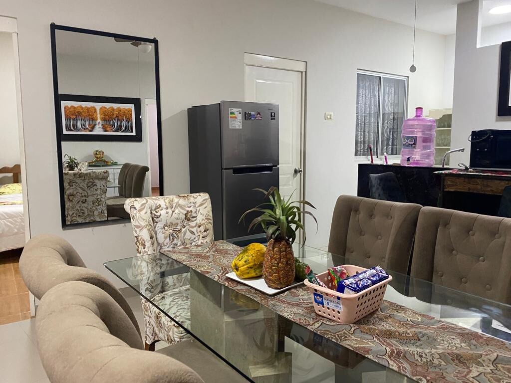 A modern dining area features a glass table surrounded by upholstered chairs. A decorative table runner showcases fresh fruit in the center, complemented by a large mirror reflecting the space. A refrigerator and kitchen appliances are visible in the background, enhancing the functional layout.