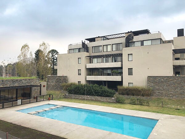 The exterior of a modern multi-story building is showcased, featuring large windows and a spacious terrace. A community pool sits in the foreground, with well-maintained grass surrounding it. Adjacent, a club house is visible, suggesting recreational options for guests.