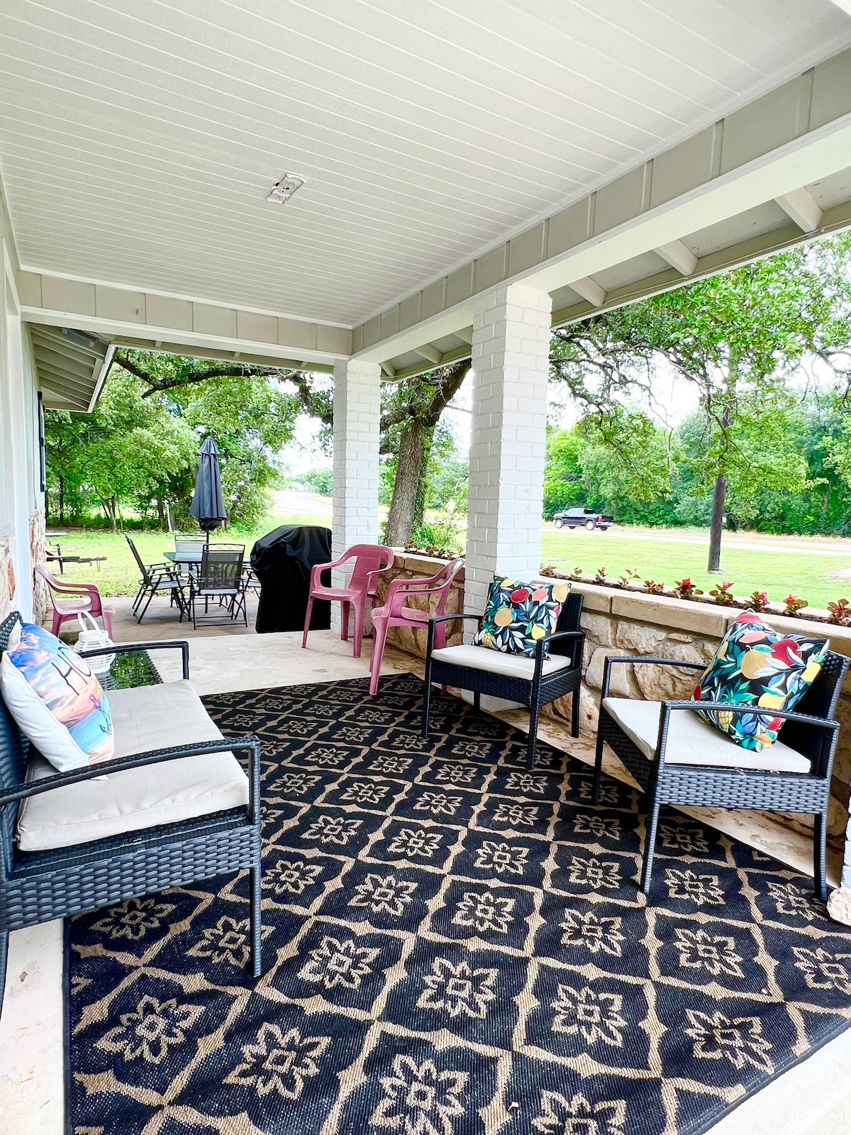 A covered porch area offers seating with two armchairs and two pink chairs, complemented by a patterned rug. A dining table is visible in the background, along with a grill, while surrounding trees enhance the outdoor atmosphere.