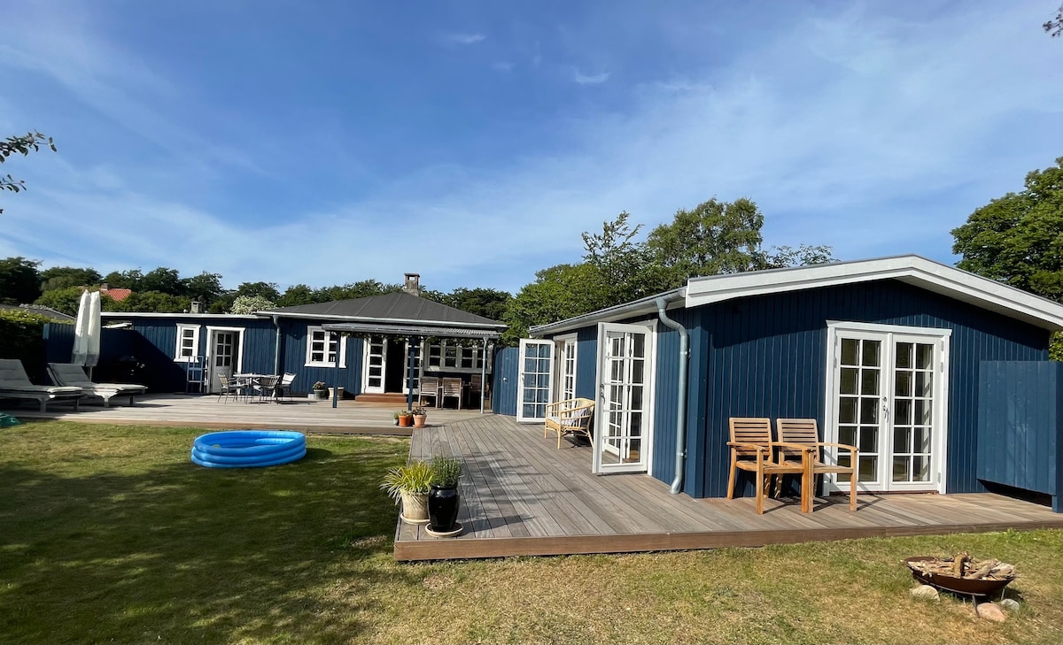 Two separate blue buildings are set on a spacious deck surrounded by greenery. A circular inflatable pool is visible on the lawn, with wooden chairs placed nearby. Large glass doors open to the main building, allowing natural light to fill the space.