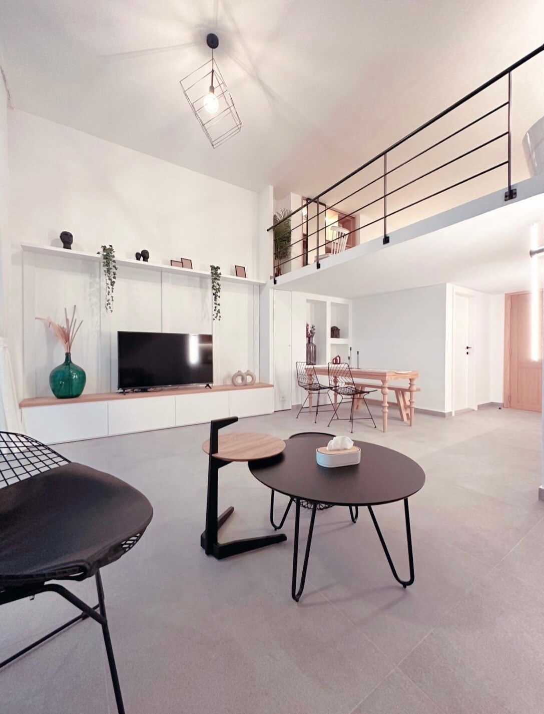 The open living area features a high ceiling with a modern light fixture. A television is mounted on a stylish white cabinet. Seating options include a black chair and a small table alongside a larger coffee table. A dining area with wooden chairs is visible in the background.