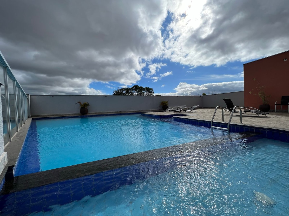 A rooftop pool is visible, surrounded by a sun terrace with lounge chairs for relaxation. Clear water reflects the cloud-covered sky above, creating a serene environment. Potted plants add a touch of greenery to the area, complementing the tranquil setting.