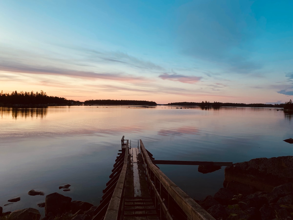 A wooden dock extends into a calm body of water, reflecting shades of blue and pink from the evening sky. Silhouettes of distant land can be seen on the horizon, surrounded by a peaceful landscape of trees and rocks.