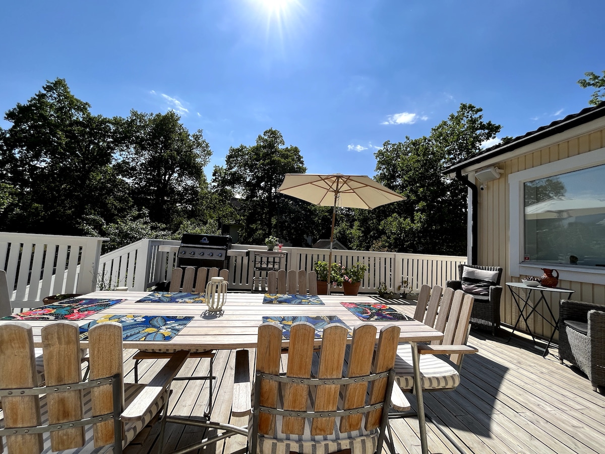 An outdoor terrace is presented, featuring a wooden dining table surrounded by several chairs. A large umbrella provides shade, while a grill sits nearby. Lush trees frame the view, with the sun shining brightly overhead, creating a welcoming space for relaxation and meals.