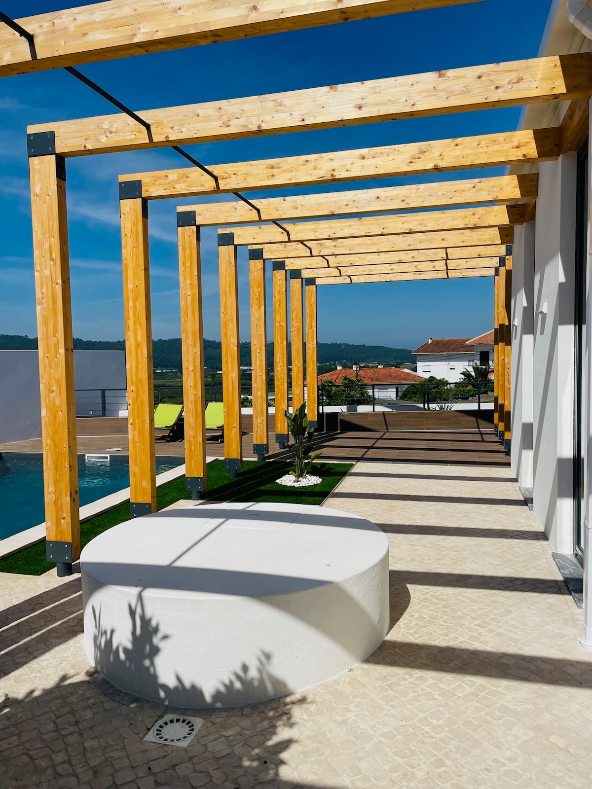 A shaded walkway features wooden beams that frame an open view of the landscape. A circular concrete bench provides a resting spot, while the surrounding area is complemented by greenery and a glimpse of the pool nearby.