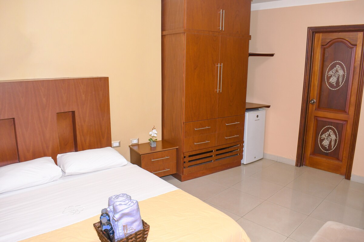 The sleeping area features a spacious bed with a light-colored blanket. A wooden wardrobe, equipped with drawers, stands against the wall. A small refrigerator is positioned next to a minimalist shelf. Neutral wall tones contribute to a calm ambiance.