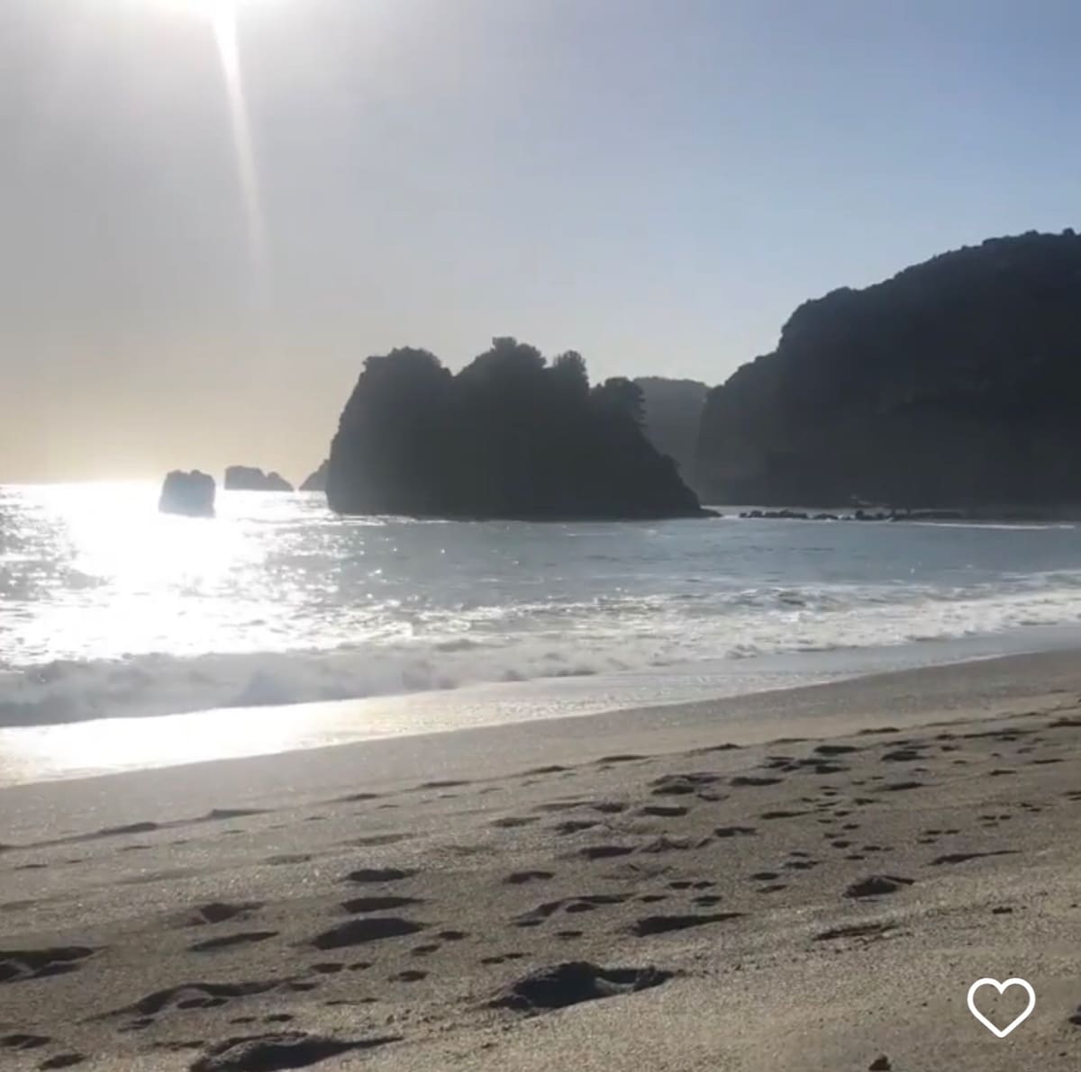 Gentle waves lap against a sandy beach, with footprints leading toward the water's edge. In the distance, rocky formations can be seen rising from the sea, framed by a soft, hazy light. The sun casts a warm glow over the tranquil scene.