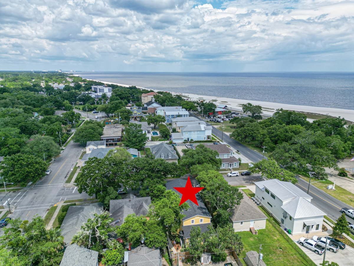 An aerial view showcases the coastal neighborhood with lush greenery surrounding a charming yellow house. The nearby beach is visible, extending along the shoreline under a partly cloudy sky. The vibrant community features a mix of structures, bordered by trees and parking spaces.