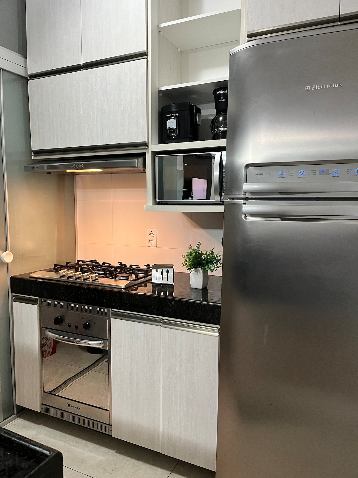 A modern kitchen is presented with sleek cabinetry and a dark countertop. Essential appliances, including a stainless steel refrigerator, microwave, and gas stove, are neatly arranged. A small plant adds a touch of greenery, while the lighting enhances the clean, organized space.