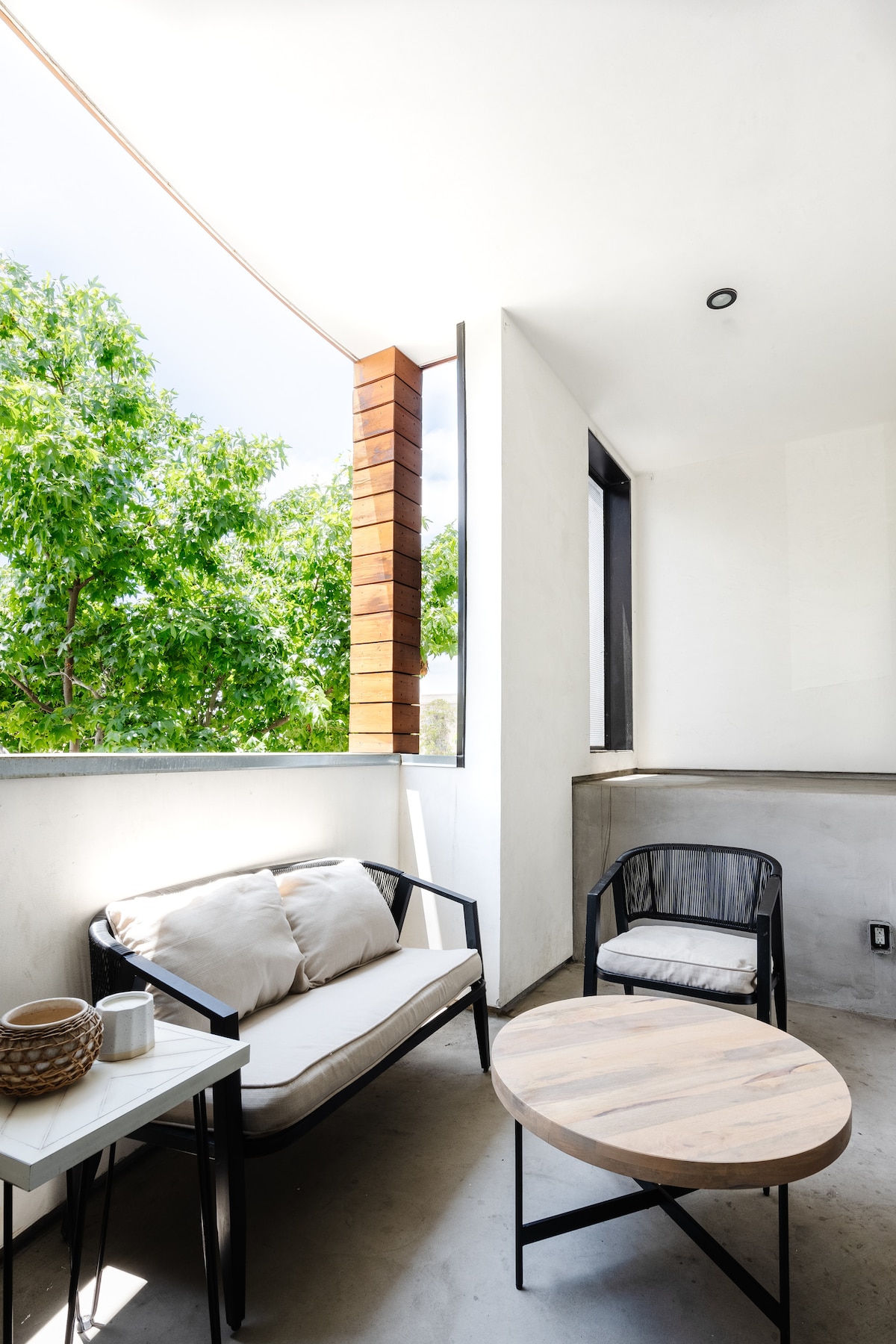 A shaded patio area features a modern seating arrangement consisting of a cushioned sofa and a single chair, complemented by a wooden coffee table. Lush greenery is visible through the nearby openings, enhancing the sense of openness and connection to the outdoors.