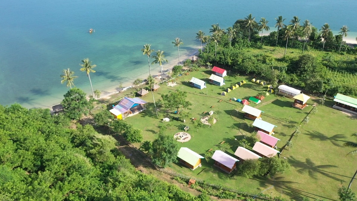 An aerial view of Beach Camp Lombok showcases a vibrant landscape of grassy areas dotted with colorful camping tents and wooden houses. The serene beach and palm trees frame the scene, with a clear blue sea in the background, highlighting the natural beauty of the location.