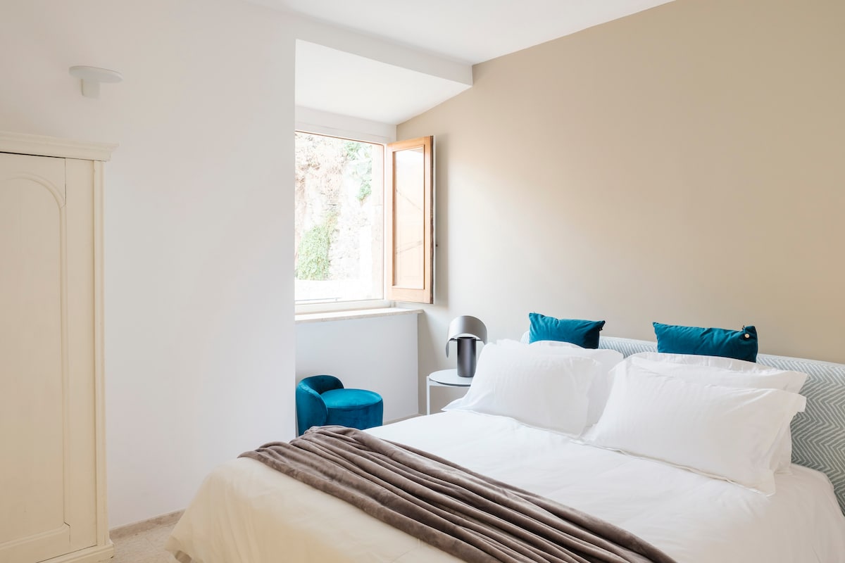 A well-lit bedroom features a spacious bed adorned with white linens and a beige throw. Blue accent pillows add color to the headboard. A small blue chair and a gray side table are positioned near a window, which is opened to allow natural light.