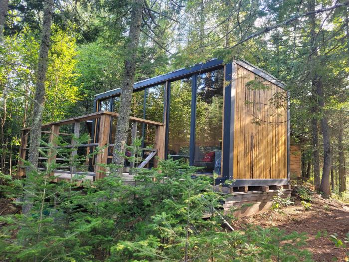 The cabin, designed with contemporary elements, is nestled among trees, featuring large glass windows that invite natural light. The exterior combines wooden siding with a modern aesthetic, complemented by a surrounding deck that offers outdoor access.