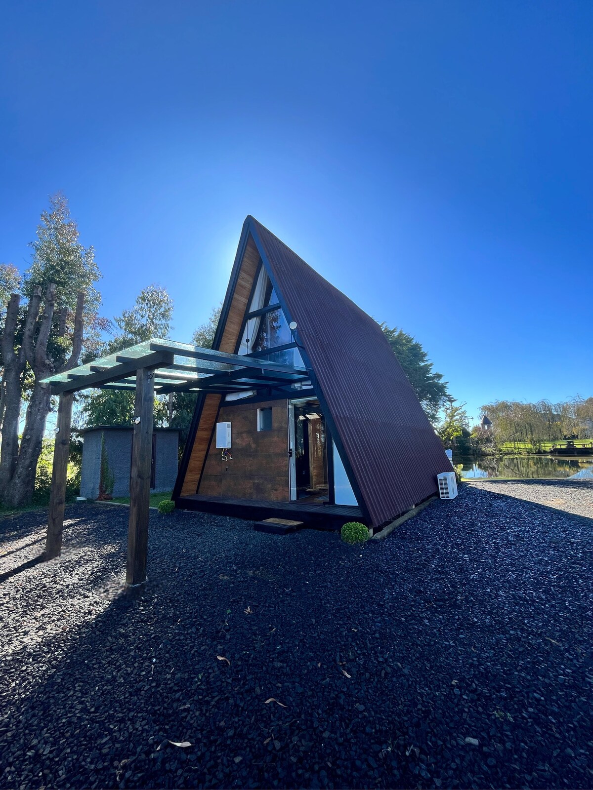 The A-frame chalet is set against a clear blue sky, showcasing its unique triangular architecture. The exterior combines wood and glass elements, framed by tall trees. A covered entrance is marked by columns, and the ground is composed of dark gravel.