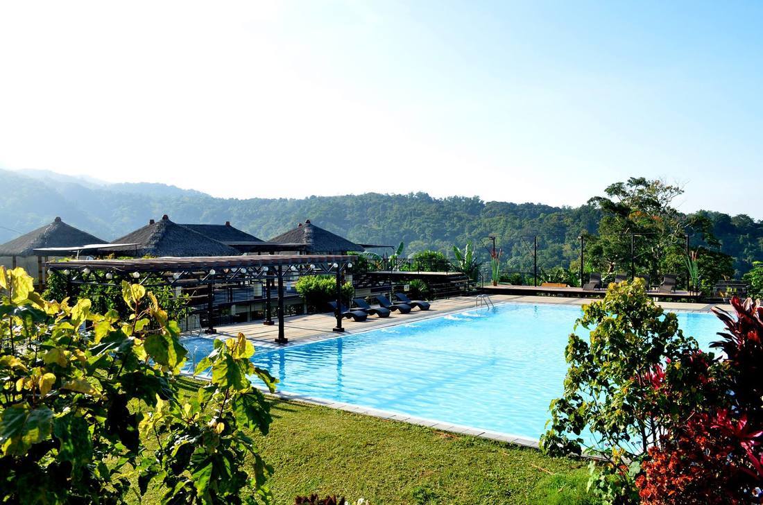 A serene outdoor pool is surrounded by lush greenery and vibrant flowering plants. The clear water reflects the blue sky and distant mountains. Thatched-roof structures provide shaded seating areas, creating a calm retreat for relaxation and enjoyment of the natural surroundings.