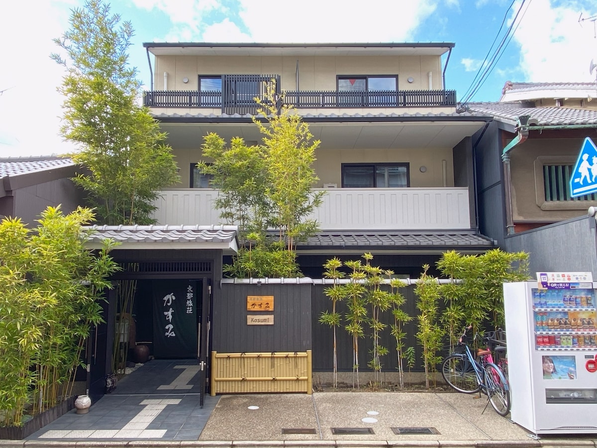 A three-story building with a traditional Japanese facade is surrounded by greenery. The entrance is marked by a black wooden gate, while bamboo plants enhance the tranquil setting. A vending machine and a bicycle are visible along the pathway.
