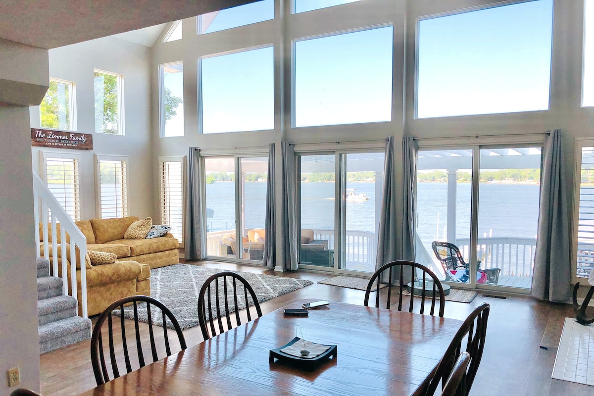 A spacious living area features large windows that provide a view of the lake. Natural light fills the room, highlighting a cozy couch and a dining table surrounded by chairs. Two sets of sliding glass doors lead to the outdoor deck.