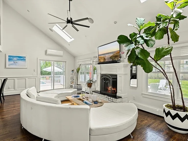 A bright living room features a curved white sectional sofa arranged around a wooden coffee table. A large television is mounted above a modern fireplace. Natural light streams in through multiple windows, while a tall potted plant adds a touch of greenery to the space.