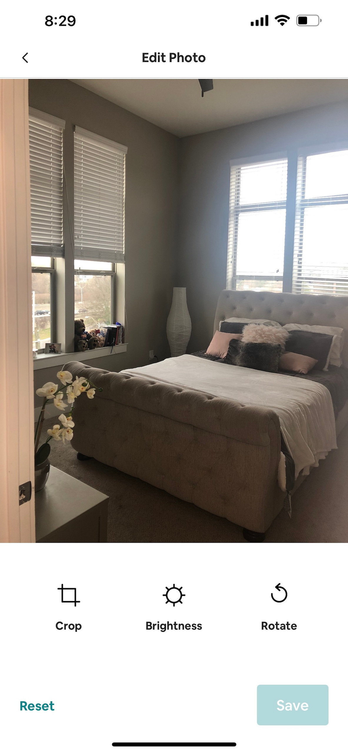 A calming bedroom features a spacious bed with light bedding and decorative pillows. Natural light enters through two large windows adorned with blinds. A stylish lamp stands beside the bed, and a small nightstand holds decorative items, enhancing the room's simplicity.