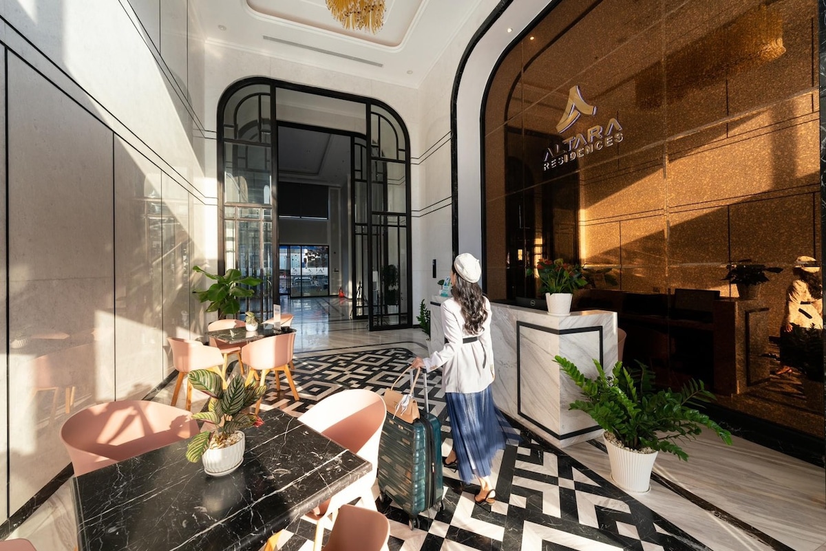 A modern lobby is viewed with a stylish floor featuring a geometric pattern. Potted plants are placed on white pedestals, enhancing the sophisticated atmosphere. A guest is seen entering, carrying luggage and dressed casually. Large glass doors lead to the exterior, inviting natural light.