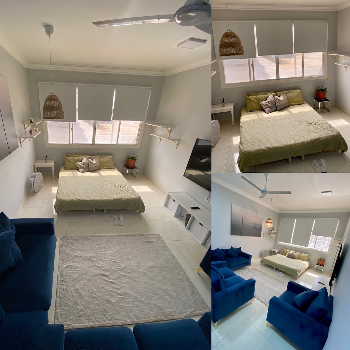 A spacious sleeping area features a large, unmade bed with earthy green bedding. Two bright windows allow natural light to fill the room. A modern television is mounted on the wall, with a cozy seating arrangement consisting of a light-colored rug and blue sofas positioned nearby.