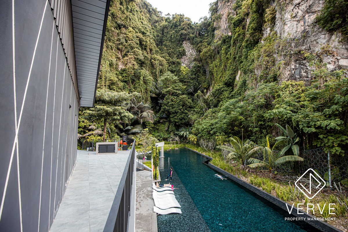 An outdoor area features a tranquil swimming pool, surrounded by lush greenery and rocky cliffs. Lounge chairs are positioned along the pool, providing a serene spot for relaxation. The modern condominium structure forms a sleek backdrop to this natural oasis.