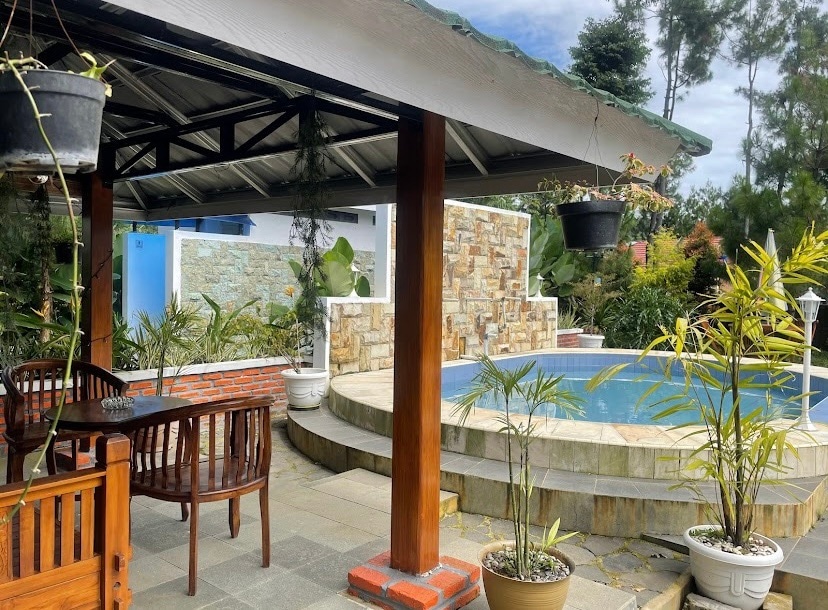 A covered outdoor seating area features a wooden table and benches. Potted plants surround the space, and a circular pool is positioned nearby, framed by stone walls. Overhead, hanging planters add greenery to the setting, creating a serene atmosphere.