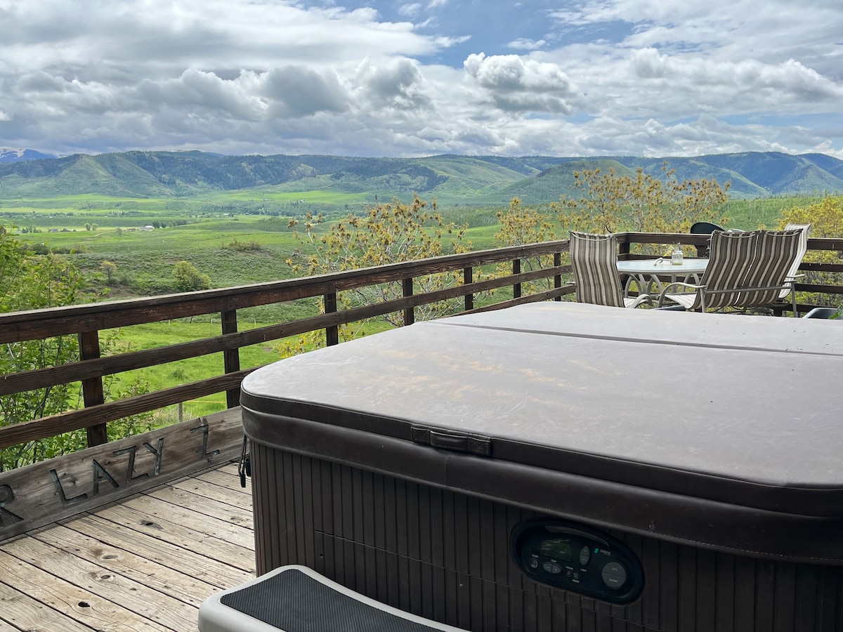 A hot tub is positioned on a wooden deck, offering expansive views of rolling green hills and the Zirkel Wilderness area in the distance. A dining table with striped chairs is also visible, accompanied by a serene sky filled with clouds.