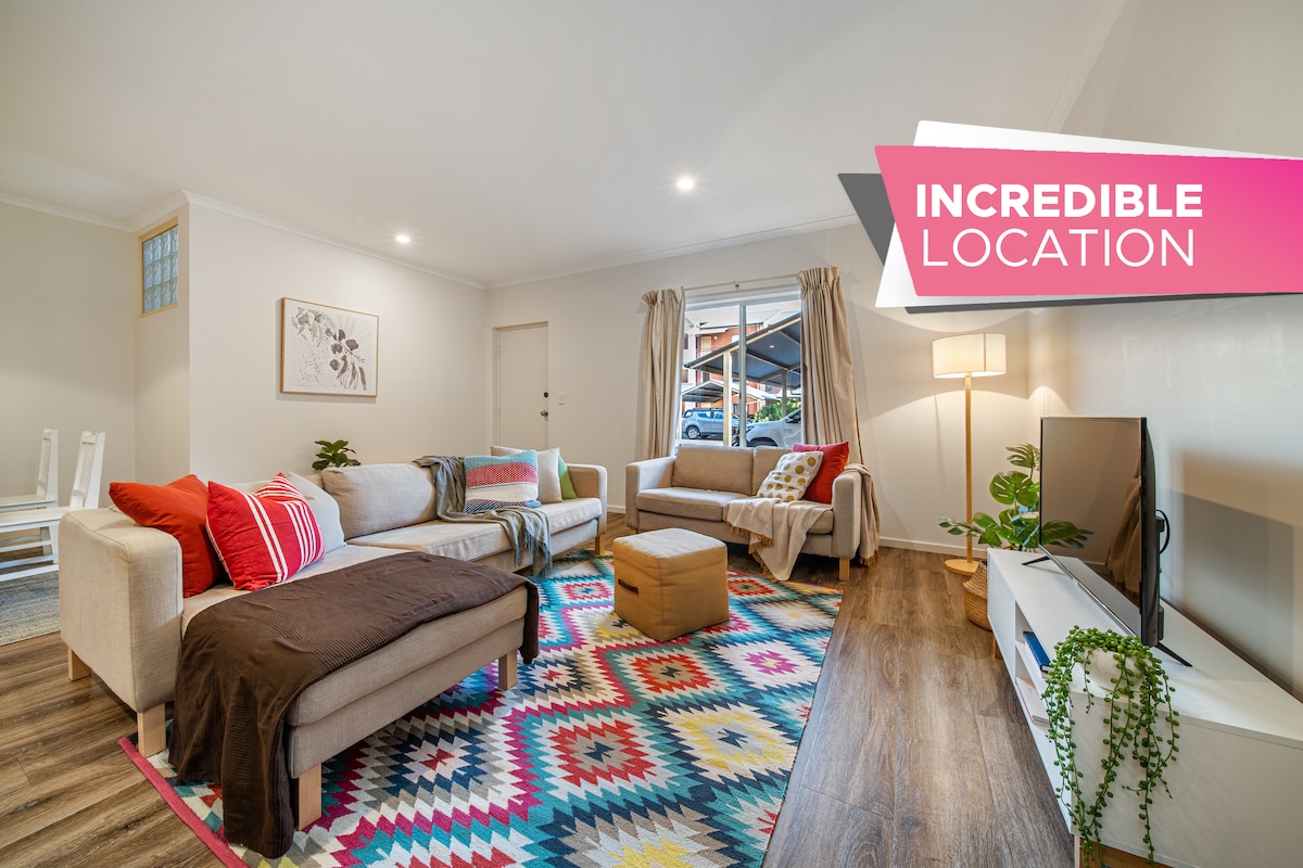 The living area displays a vibrant, patterned rug covering the floor beneath a comfortable three-seater chaise lounge and a two-seater lounge. A coffee table sits centrally, with accessories around the room, including a floor lamp and a TV on an entertainment unit. Natural light filters in through a window.
