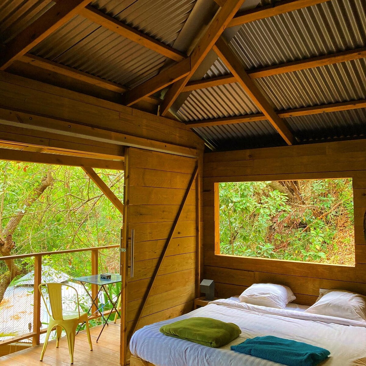 A cozy sleeping area features a spacious bed with white linens and colorful towels. Light wooden walls create a natural ambiance, while a large window offers views of lush greenery. A small round table and chairs are positioned nearby, enhancing the serene setting.