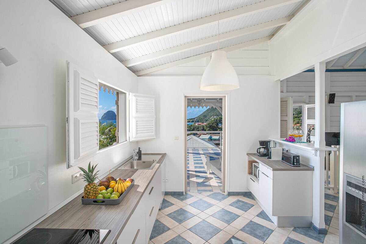 A bright and airy kitchen features white cabinetry and a large window offering a view of the Caribbean Sea. A countertop displays an array of fresh fruit, complemented by a contemporary light pendant overhead. The tiled floor adds a structured element to the open space.