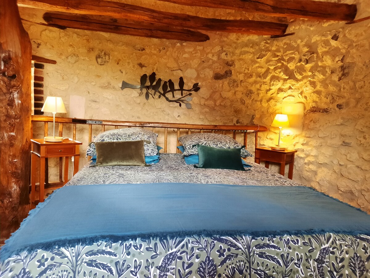 A spacious bedroom features a stone wall with exposed wooden beams. Two bedside tables with lamps flank a large bed adorned with a blue throw and patterned bedding. Soft lighting enhances the cozy ambiance of the room.