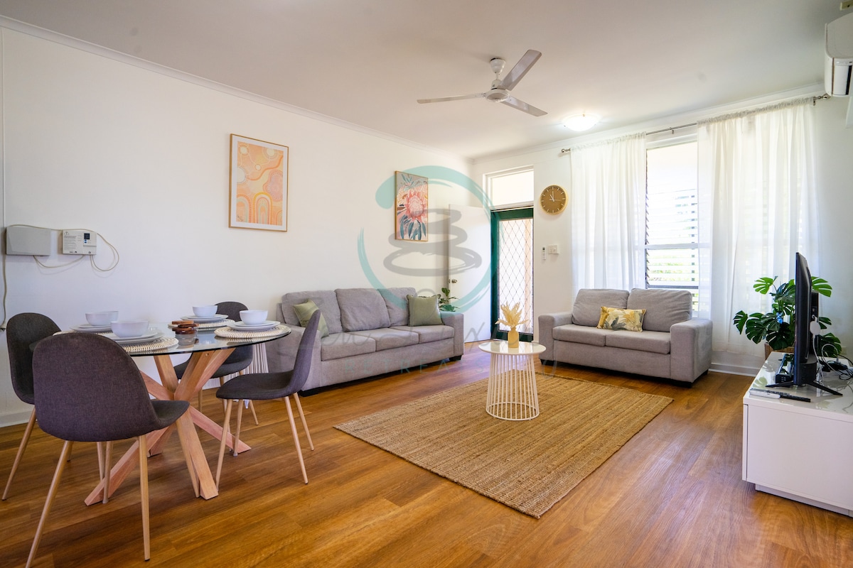 A modern living area is presented with light wood flooring and neutral-toned furnishings. A round dining table with matching chairs is arranged beside a cozy sofa. Two large windows allow natural light to brighten the space, complemented by tasteful artwork on the walls.