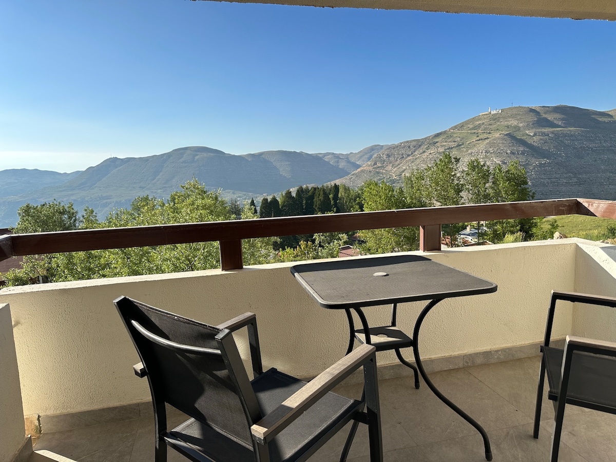 A private balcony features a table and two chairs overlooking a scenic mountainous landscape. The view includes rolling hills and trees under a clear blue sky, providing a peaceful outdoor space for relaxation.