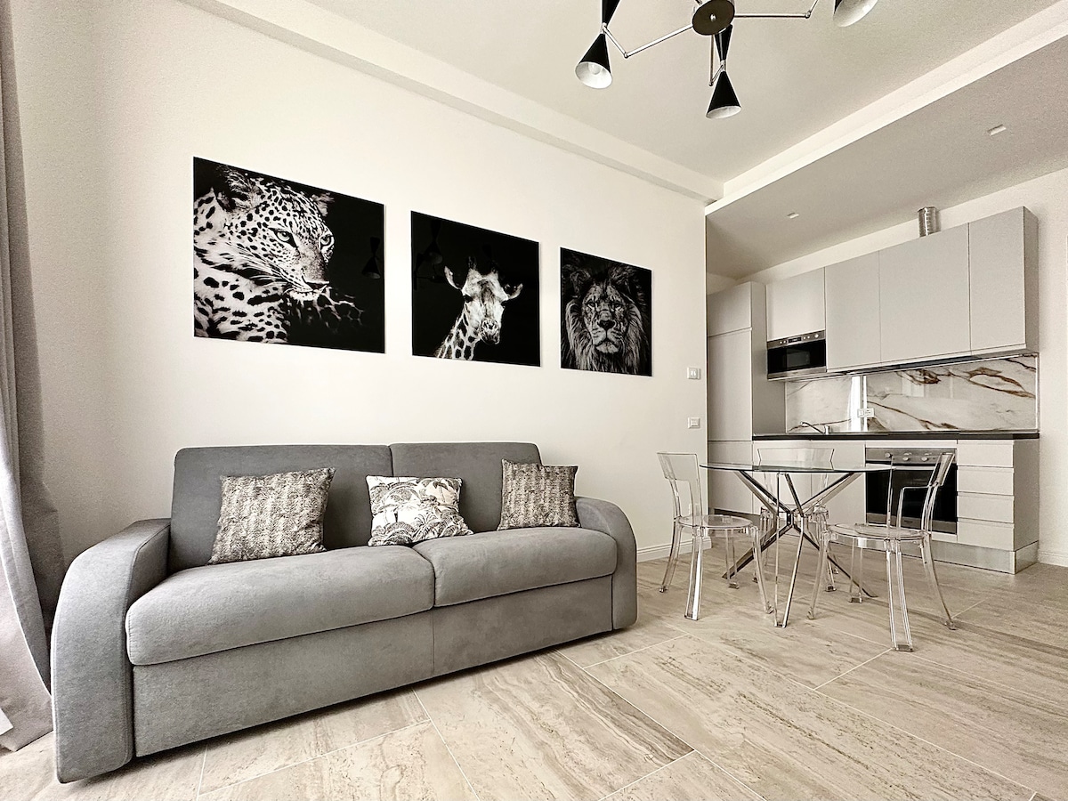 A modern living space features a gray sofa adorned with patterned cushions. Three black-and-white animal portraits are displayed on the wall. A glass dining table surrounded by transparent chairs complements the sleek kitchen area in the background.
