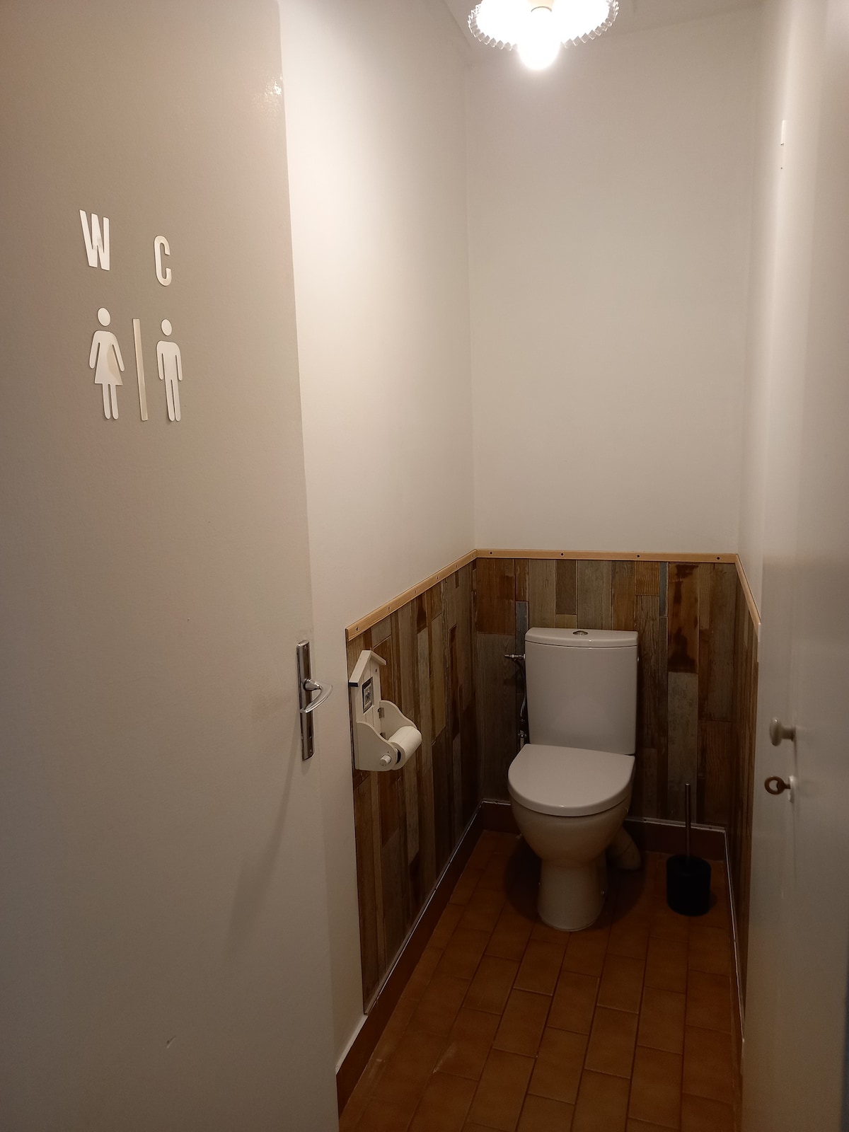 A compact restroom features a modern toilet positioned against a wooden accent wall. The space includes a toilet paper holder and a small waste bin. Light is provided by a ceiling fixture, creating a well-lit atmosphere.