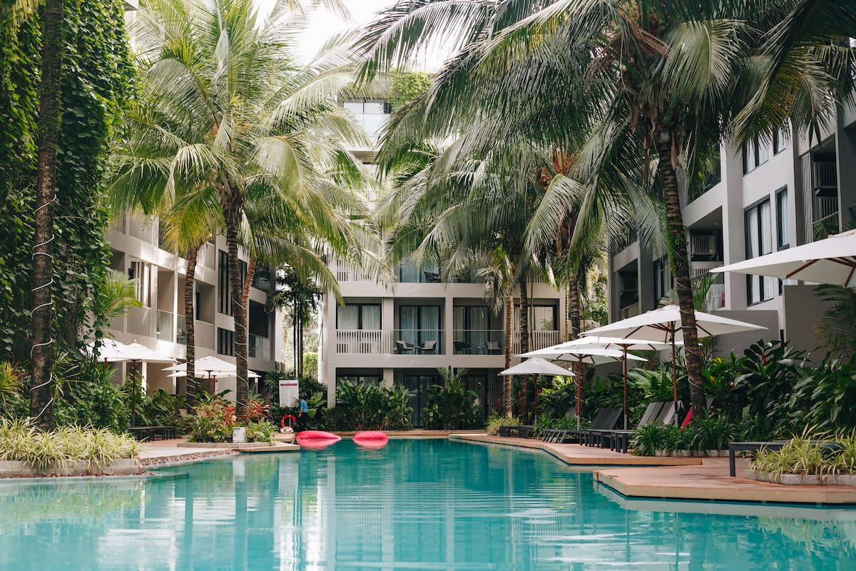 A serene swimming pool is surrounded by lush palm trees and tropical plants, with sun loungers positioned under large umbrellas. Two kayaks float gently on the water's surface, reflecting the greenery and inviting relaxation in a vibrant setting.
