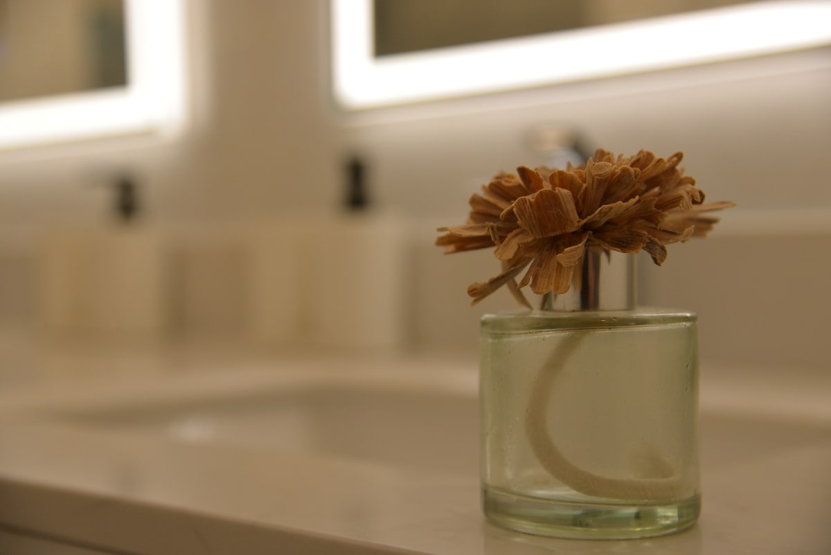 A bathroom counter features a delicate vase holding a dry flower arrangement. The vase rests on a smooth marble surface, complementing the contemporary design. Soft lighting from illuminated mirrors enhances the clean lines and simple decor of the space.