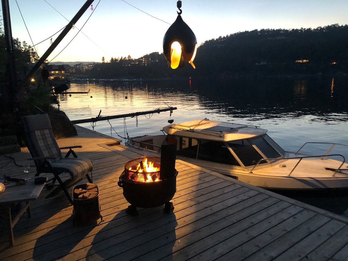The image captures an evening scene by the waterfront, featuring a cozy fire pit with flames, surrounded by a wooden deck. A comfortable chair is positioned nearby, and a boat is moored close to the shore, reflecting the soft light of the surroundings.